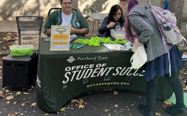 Two people, Noah and Alice sitting at a table with an Office of Student Success tablecloth. They are helping a student, who has their back turned and is picking up an item from the table.