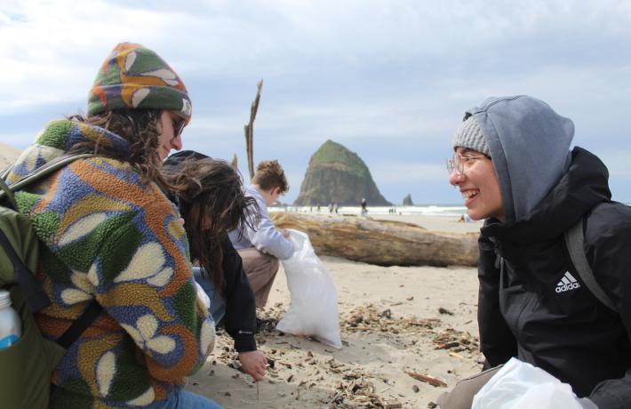 Two people are seen smiling on the beach and looking at the sand with a large rock in the ocean in the background.