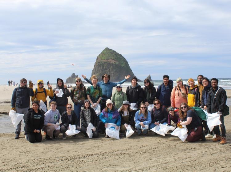 A group of people is shown standing in front of the ocean and a large rock. The students in the front row are holding trash bags.