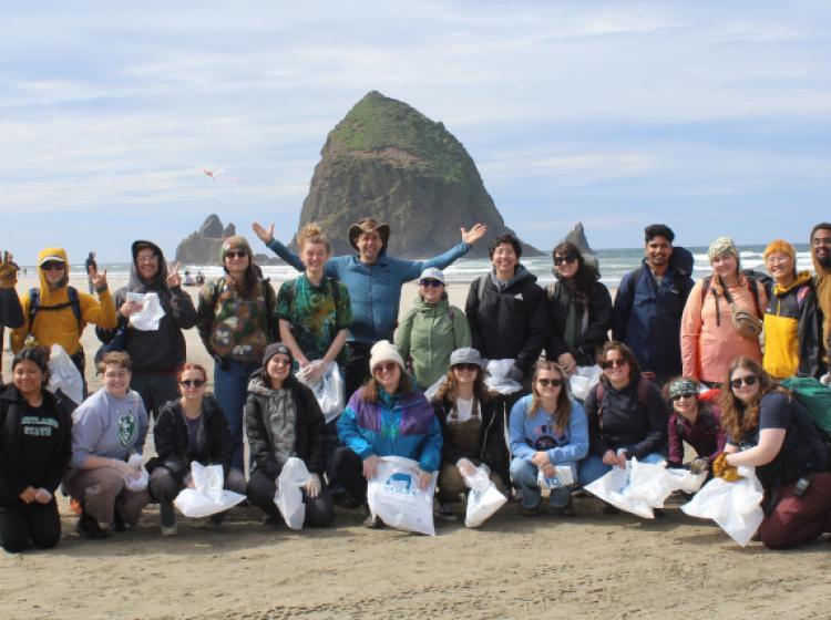 2 dozen people holding filled garbage bags from a litter cleanup taking a group photo on the beach in front of Haystack Rock