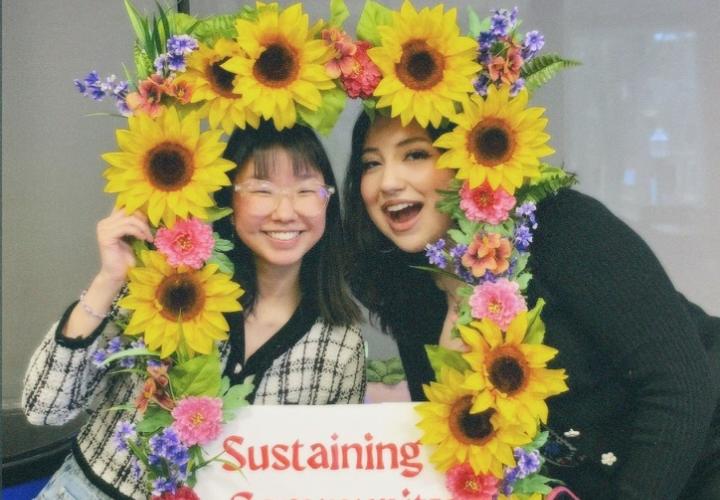 Two students smiling and holding a sunflower picture frame with the words "sustaining community"
