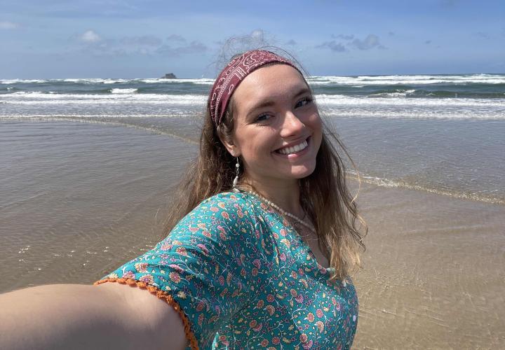 A person with blonde hair is shown smiling on the beach in front of the ocean.