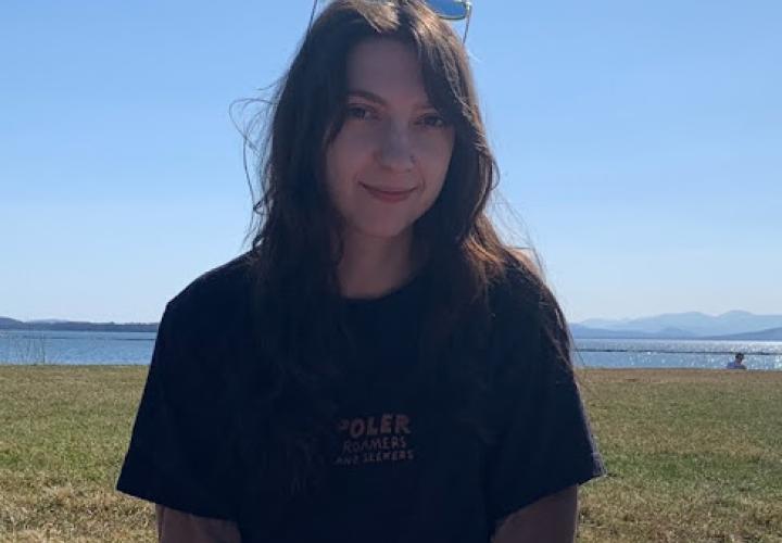 A person with long brown hair and sunglasses on their head is shown standing in front of a field and a body of water.