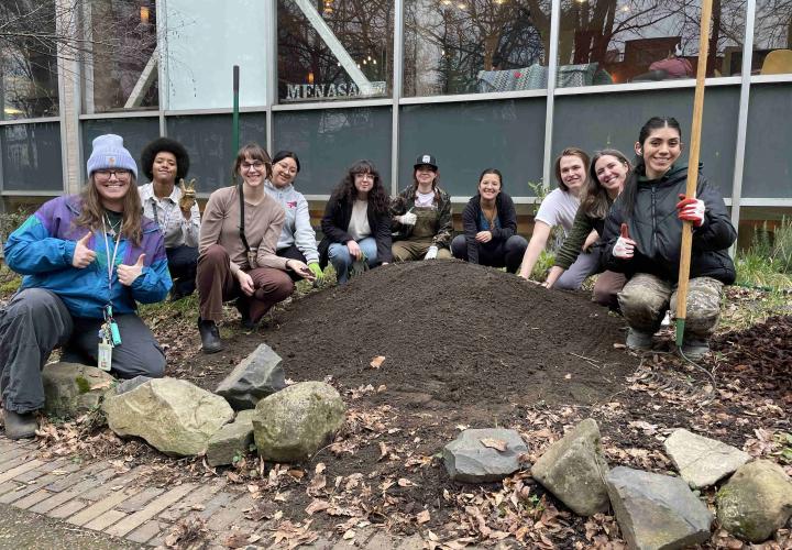 A group of people next to a mound of dirt is shown. They are crouching with garden tools and smiling.