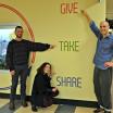 Three people pointing to wall mounted sign saying give take share