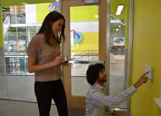 one student standing overlooks and takes notes while another student looks at sensor on the wall