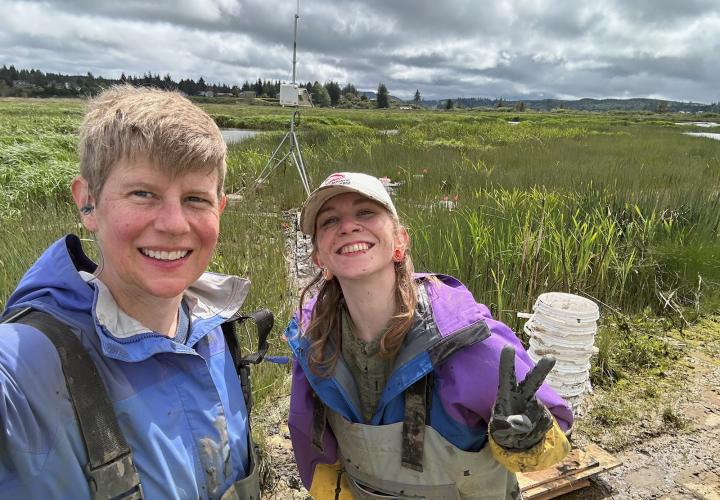 Two female scientists stand on the edge of a green wetland, smiling