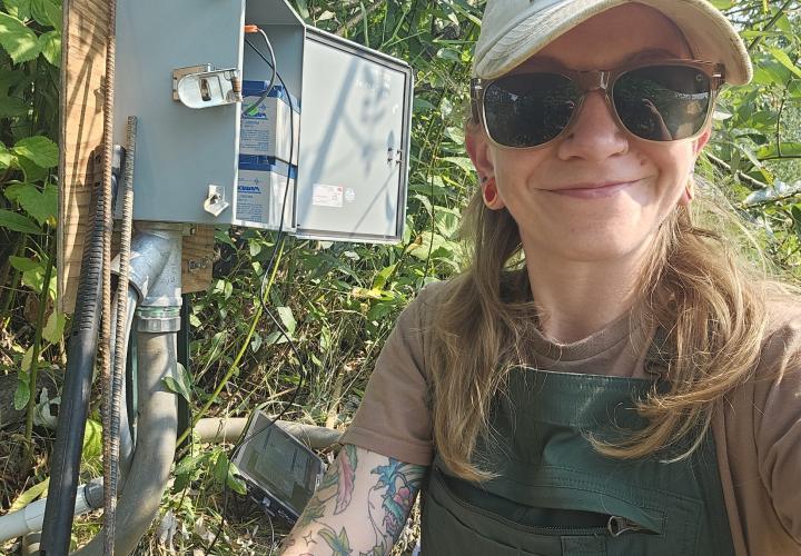 A woman wearing sunglasses and a hat smiles sitting next to a USGS gage box that is connected to a laptop.