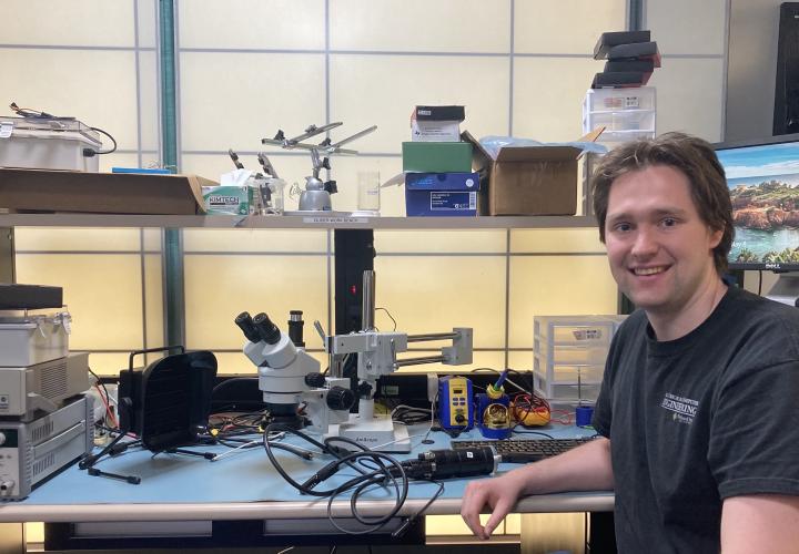 A white man sits at a lab bench surrounded by a microscope and electronic equipment