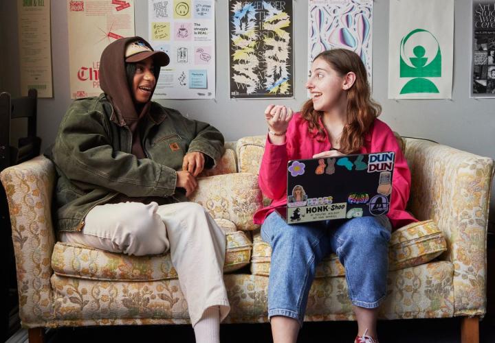 two people sitting on a tan couch talking in front of various posters on the wall