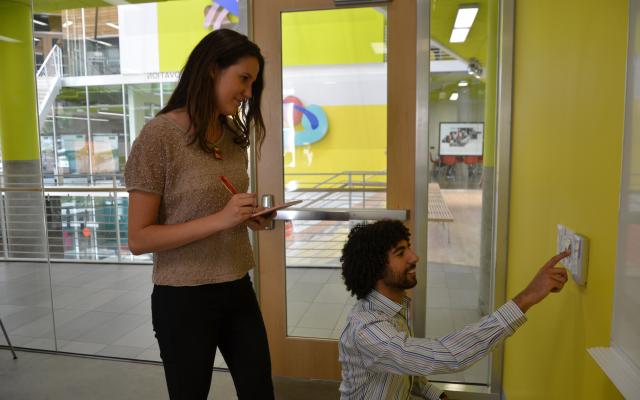 one student standing overlooks and takes notes while another student looks at sensor on the wall