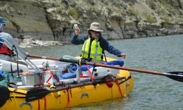 A person sits near the back of a yellow raft on a river with equipment strapped down on the boat, holding the end of a line that is tied to a measuring device in the water