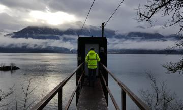 USGS field researcher standing in front of small concrete room overlooking lake with mountain in the background