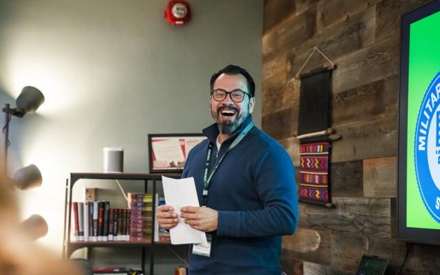 Ron Mansolilli smiles while holding a document in front of a screen displaying the Military Friendly Gold School seal