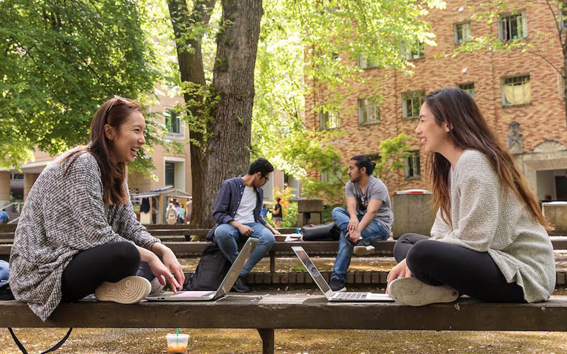 Two female students sitting across from each other outside, talking and working on their laptops