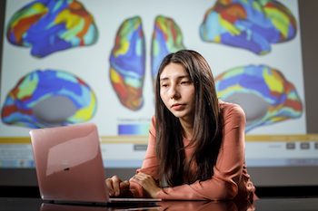 a person seated at a table working at a laptop. Behind them is projected colorgul digital renderings of a brain