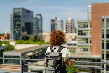 a student wearing a back pack stands at a balcony railing with the city of Portland in the back ground. 