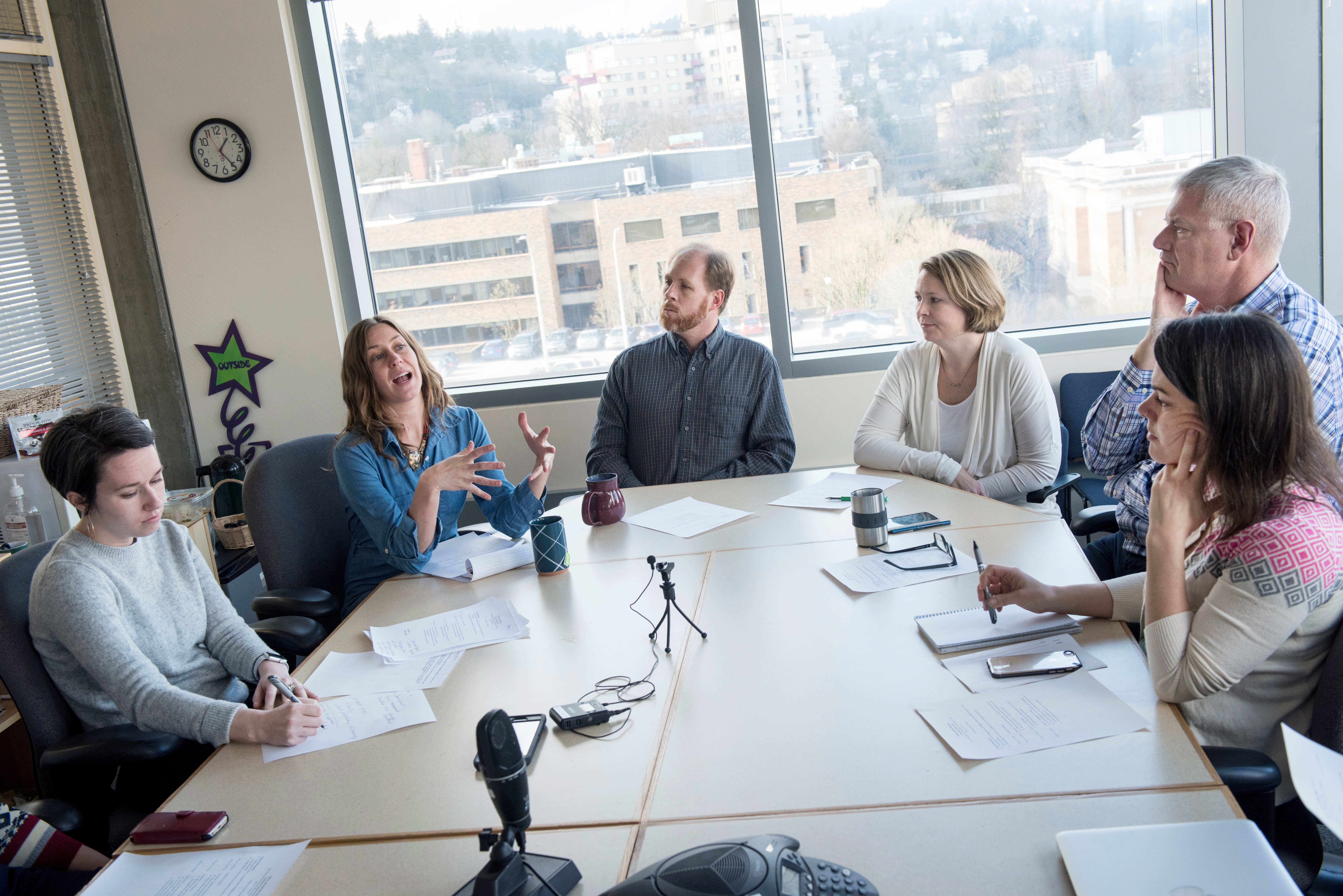 six people seateda round a conference table. A woman is speaking and all other participants are actively listeining to her