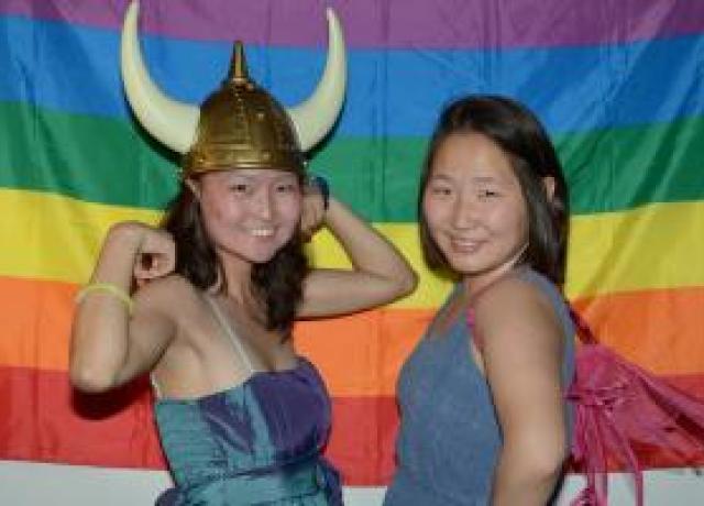 Two students standing in front of a LGBTQ rainbow pride flag. One student wears a viking helmet.