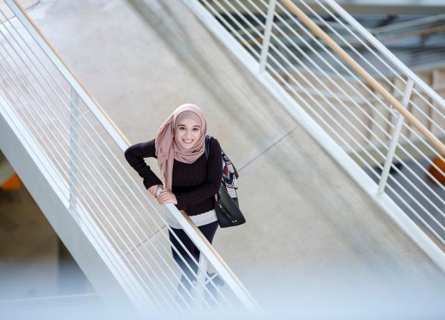overhead view of a woman wearing a head scarf,  standing on a bridge, looking up at the camera. 