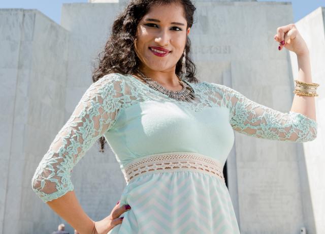 Woman in an aqua dress standing in front of the Oregon Capital Building, flexing her bicep in a show of strength and women's solidarity