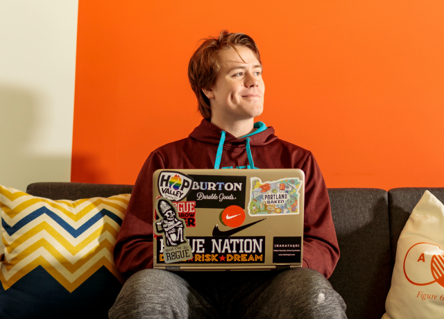 student seated on a couch in front of an orange wall, with their laptop opened. 