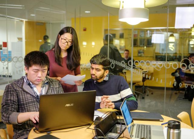 Three students around a table looking at a laptop. One student appears to be typing