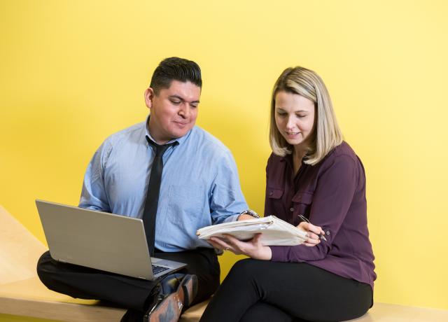 two individuals seated on a bench, reviewing information in a spiral notebook.