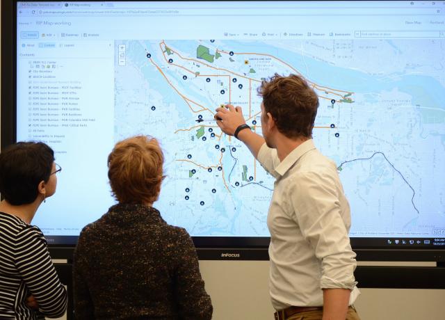 three individuals are looking at a large monitor showing a map with various data points