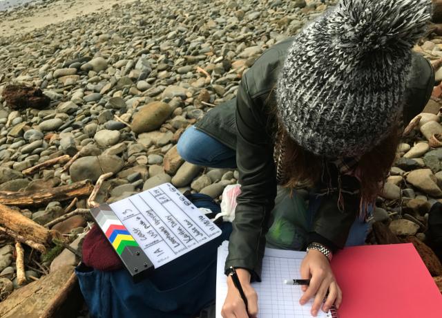 looking down at a person seated on a rocky patch of ground. They write in a notebook and a film clapper sits on a backpack next to them.