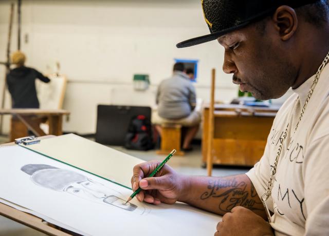 a student sits at a table creating a portrait in pencil