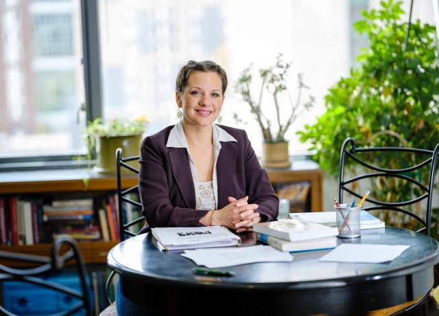 a woman in a business suit sits at a table with stacks of books and paper on the table