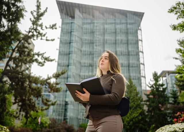 A PERSON HOLDING AN PENBOOK STANDS OUTSIDE IN A CITY PARK WITH A BUILDING INTHE BACKGROUND