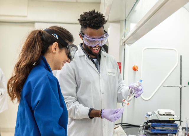 Two students in lab coats with safety glasses are manipulating a beaker with a clear liquid