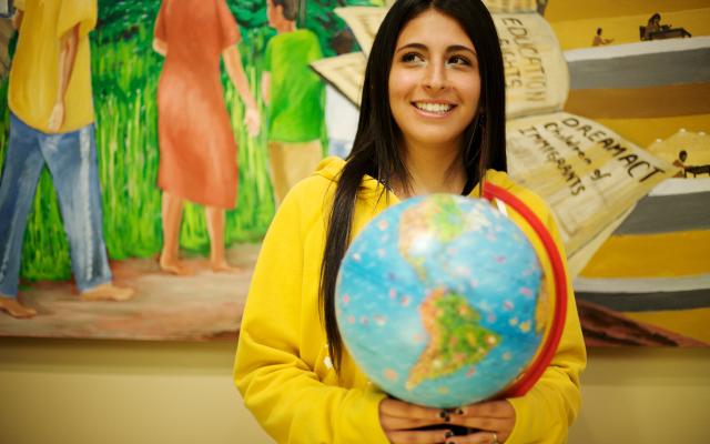 student holding a globe stands in front of a mural
