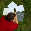 student in a red shirt, laying on their stomach in the Park Blocks grass, textbooks and notebooks are spread out in front of them.