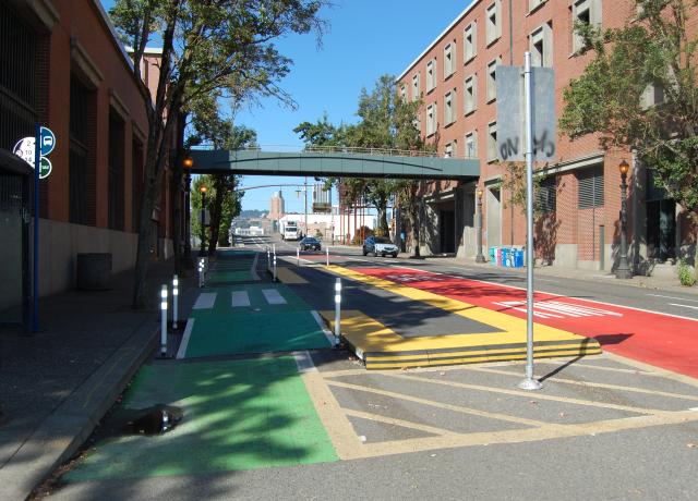 A red-painted bus lane in Portland, Oregon, with the words "BUS ONLY" in white, runs parallel to a green-painted bike lane. There is a buffer lane and bus stop in between. In the background, a pedestrian skybridge offers an elevated way to cross the street.