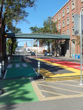 A red-painted bus lane in Portland, Oregon, with the words "BUS ONLY" in white, runs parallel to a green-painted bike lane. There is a buffer lane and bus stop in between. In the background, a pedestrian skybridge offers an elevated way to cross the street.
