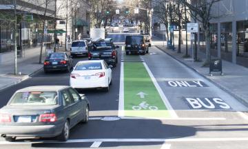 Cars drive on a street in Portland, Oregon that also has a bus-only lane and a bike lane. A van crosses the bike lane ahead in order to make a right turn.