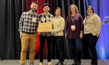 Three members of the student team stand smiling with their award from the OAPA and representatives from the OAPA.