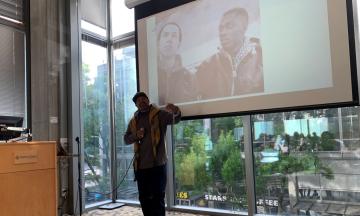 Mic Crenshaw stands with his arm raised in front of an image of his two friends from the 70s.