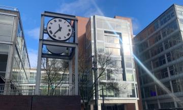 An black-and-white clock reading 11:36 stands to the left of the glass windows of the College of Urban and Public Affairs.