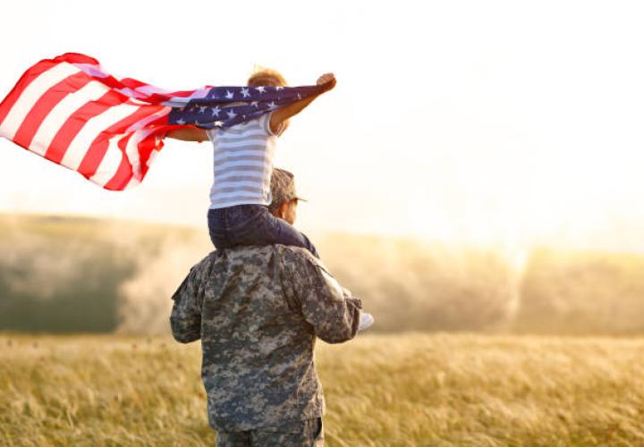 a child holding a waving american flag sits on a man's back. the man is in army uniform, both have backs turned to camera