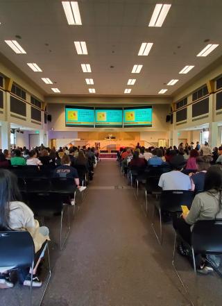 A pathway cuts through the middle of two sections of event attendees. Attendees are seated in many rows facing the stage in the front of the room. The pathway leads directly to the stage with three bright teal projector screens above the podium.
